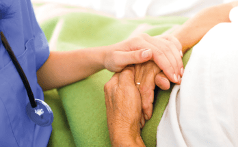 Ärztin in blauem Gewand hält die Hände einer Patientin in weißem Gewand, die mit einer hellgrünen Decke zugedeckt ist.