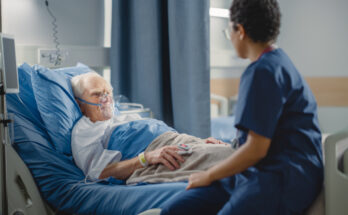 Hospital Ward: Friendly Head Nurse Connects Finger Heart Rate Monitor or Pulse Oximeter to Elderly Patient Wearing Oxygen Mask Resting in Bed. Nurse Does Patient Checkup After Successful Surgery