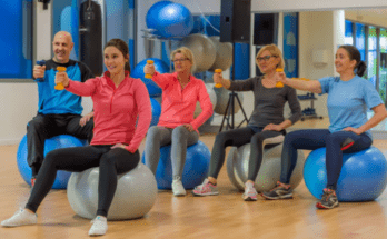 5 Personen in Sportkleidung sitzen auf Gymnastikbällen und halten mit ihrer rechten Hand eine Hantel vor sich