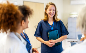 Happy nurse holding a tablet and smiling while engaging in a briefing with the medical team in a bustling hospital corridor