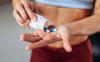 Close up of a fit, muscular sportswoman in workout attire pours blue and white supplement capsules into her palm, emphasizing fitness, health, and active living